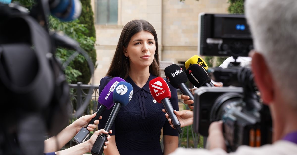 A spokesperson engages with media during an outdoor press interview surrounded by microphones.